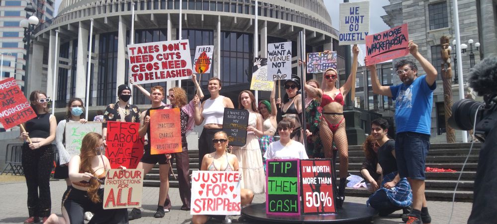 Fired Up Stilettos representatives and their allies on the steps of the New Zealand Parliament, calling for the nation's politicians to recognize its petition to improve strippers working conditions in Aotearoa. The workers rights activists hold signs that read 'Strippers Rights Are Workers Rights'; 'Labour Protections For All'; 'For-Fucks-Sake Labour - End Wage Theft'; 'Taxes Don't Need Cuts CEOs Need To Be Leashed'; 'Your Inaction Enables Exploitation'; 'Justice For Strippers'; 'Tip Them Cash'; 'I'm Worth More Than 50%'; 'I Want To Strip Off My Clothes Not My Rights'; 'We Want A Venue Not A Pimp'; 'Respect Strippers'; 'We Won't Go Quietly'; and 'No More Whorephobia'.
