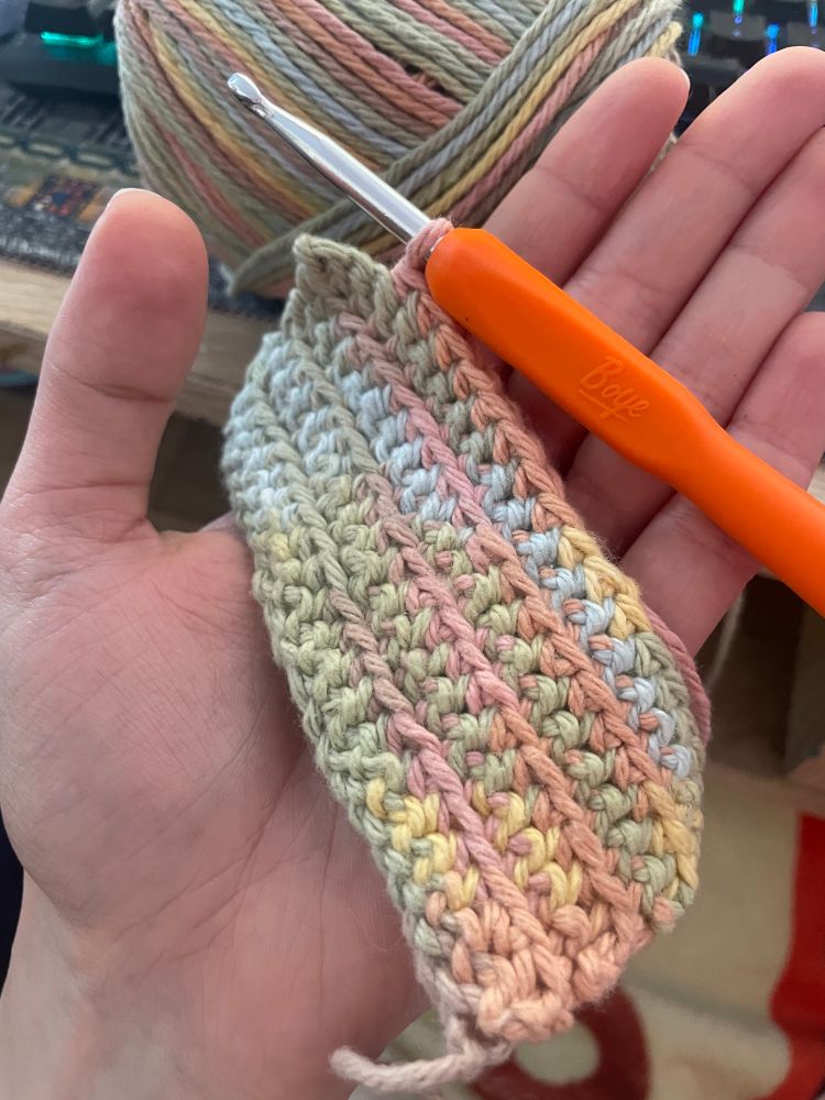 A splayed hand holding a half finished  pastel rainbow crocheted washcloth, along with the five mm hook being used to make it and a loop of yarn still attached.