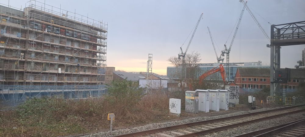 New buildings in partial states of build, with gangly cranes poised in silence. Colourful houses fringe the top of Totterdown with glowering sky behind. 