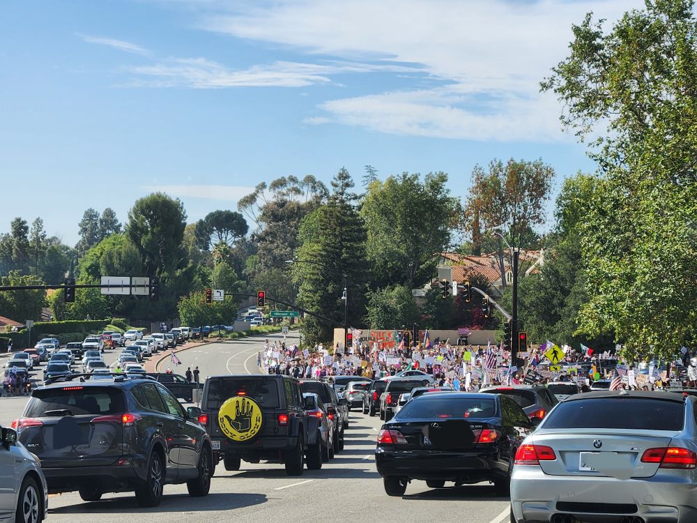 Thousand Oaks No Kings Protest at the intersection of Lynn and Hillcrest, with huge crowds of people carrying signs and flags surrounding the intersection while approaching traffic is heavily backed up in both directions. 
