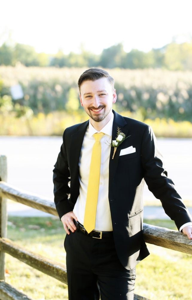 A man in a black suit leaning against a wooden fence with a field of flowers behind him.