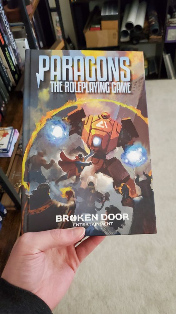 A hand holding a book titled "Paragons: The Roleplaying Game" in front of a bookshelf. The book's cover depicts a group of characters fighting a large robot.