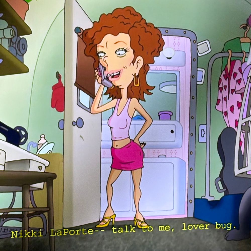 Red-head lady named Nikki LaPorte standing in her trailer home while taking on the phone. She’s wearing a pink tank top and skirt, and yellow high heels. 