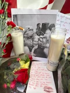 Two candles frame a photo of David Lynch being kissed on the cheek. In front of this there are flowers and a handwritten note with a lipstick kiss mark on it.