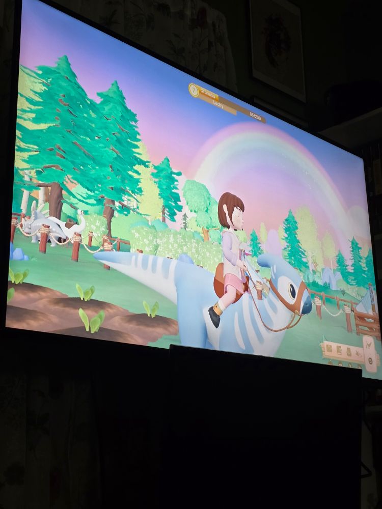 A picture of Paleo pines played on a TV, a girl with short brown hair and bangs sits on a light blue dinosaur with crops behind her and two other dinosaurs in the distance. Above her is a purple and blue evening sky, a rainbow arcing beautifully across it. 