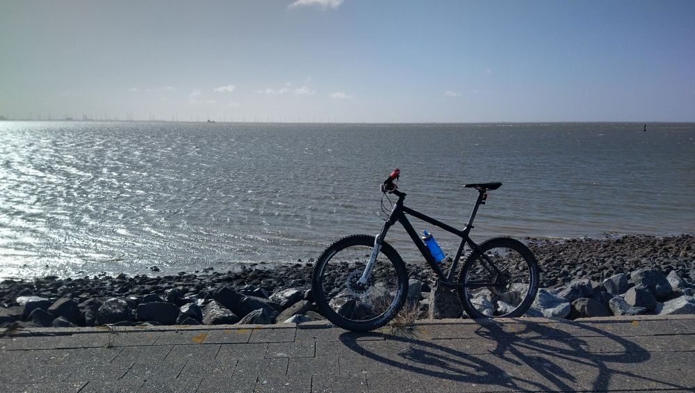 Fahrrad an Wellenbrechersteinen vorm Meer. Im Vordergrund Asphalt, das Fahrrad, dahinter schroffe, große Steine dann das Meer, was fast in das Grau des Himmels übergeht.
Das Fahrrad ist ein Mountainbike in Schwarz mit einer Wasserflasche im Rahmen 