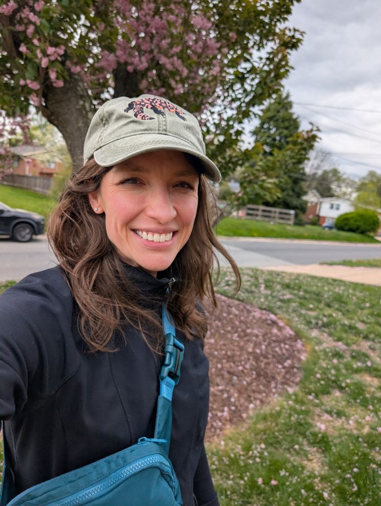 Smiling woman with brown hair, black jacket, green purse with a green baseball hat with a Gila monster on it, standing in front of a cherry tree covered in pink blossoms. 