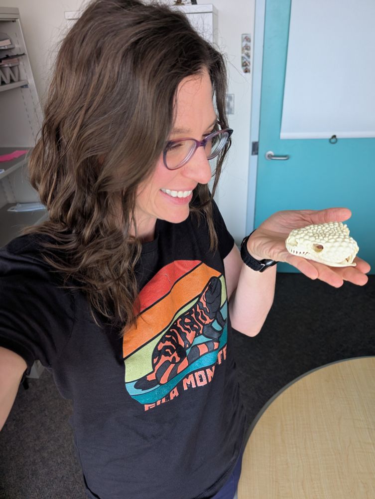 Dr. Melissa Wilson, wearing a black shirt with a Gila monster on it and purplish glasses, holding a replica of a Gila monster skull and smiling. 