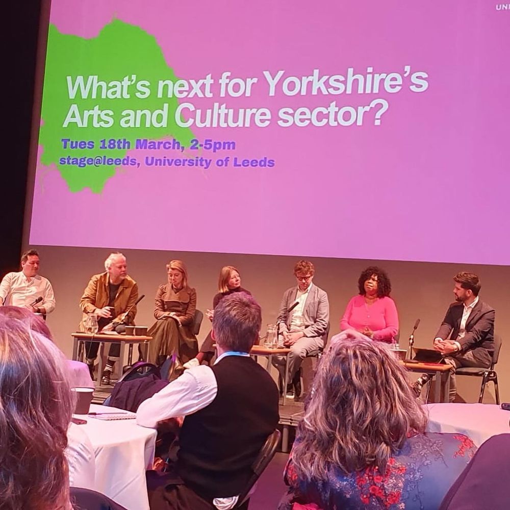 seven panelists sat on chairs on a stage with a bright pink screen behind reading What’s Next For Yorkshire’s Arts & Culture Sector
