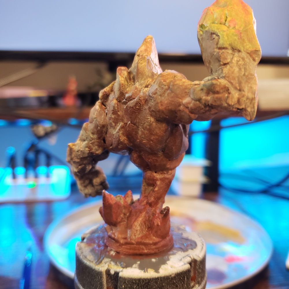 A miniature of a Earth elemental in mid layers of painting, with vibrant yellows and pinks on its upraised right arm. A lower view of the back.
