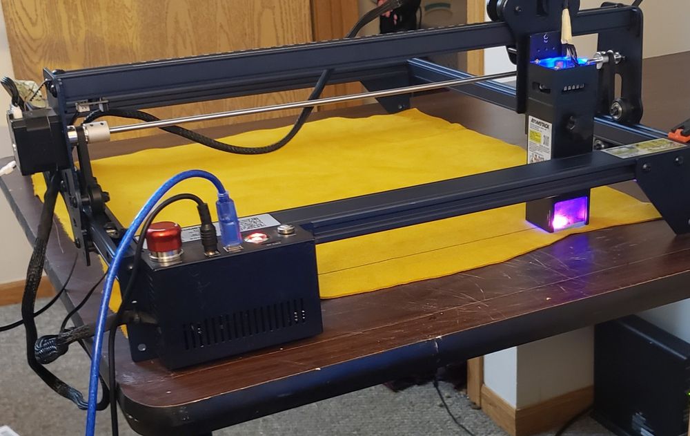 An Atomstack A10 laser engraver on a cheap, school reminiscent table, its laser bright from burning, over a large patch of vibrantly yellow suede.
