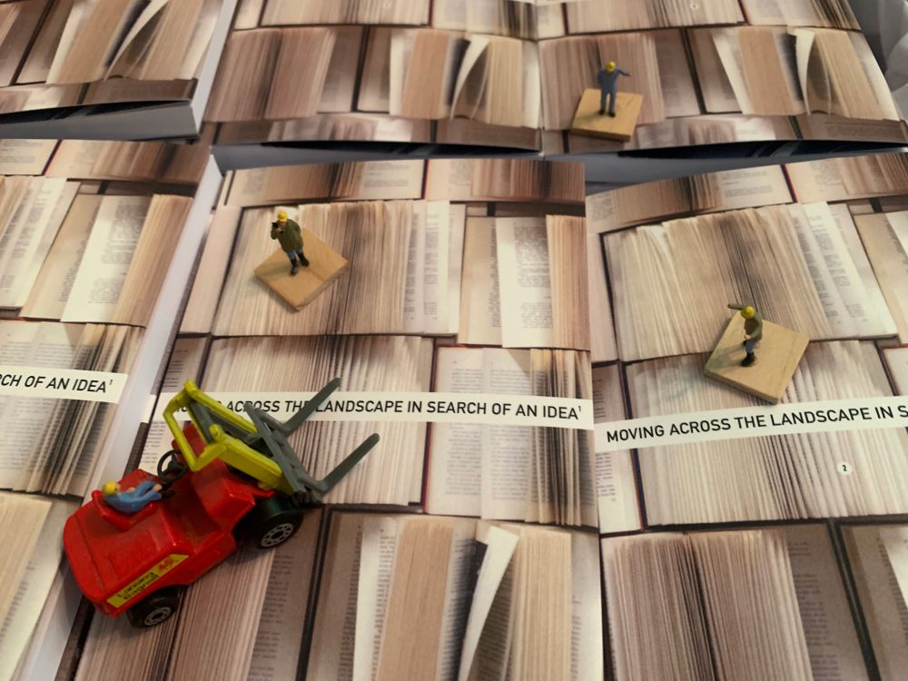 A tiny forklift and HO scale warehouse figures are standing on a plain of books as if they are sized for reality and the books are just extra huge.