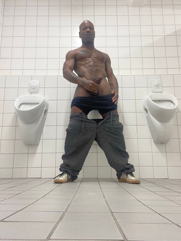 Bishop stands in front of a urinal, pants down, dick out. They look to the camera