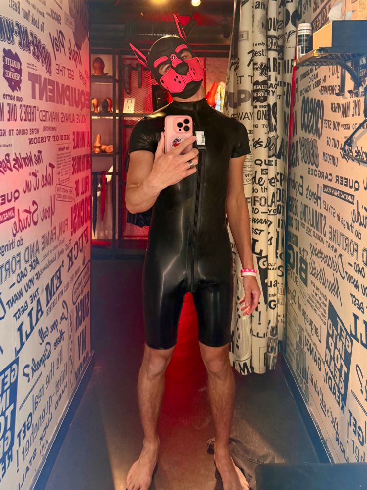 I’m in a changing room, wearing a black rubber surf suit, holding my phone up to take photo from mirror reflection, and wearing a pink pup hood. 
