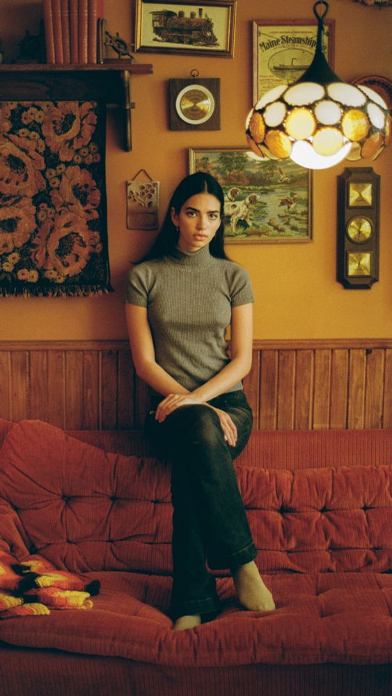 Portrait of a woman sitting on the top of the couch. She is in a 70s decorated room. The photo feels very warm due to the film being used as well as the overall room. 
