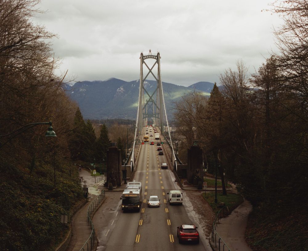Medium format film photo of the lions gate bridge in Vancouver. Shows cars driving across a bridge with mountains in the distance and trees lining the side 