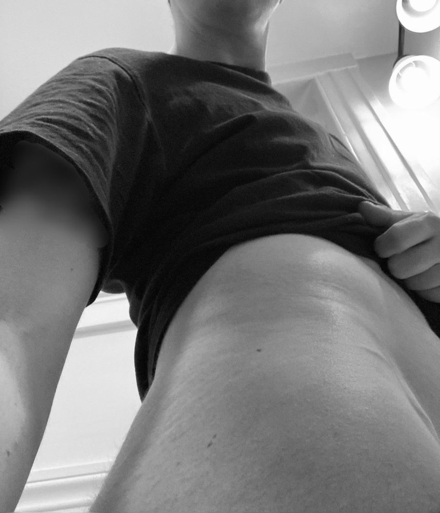Black & white photo of Cedar as if they are holding your head next to their thigh and looking up at their hip, midriff, & where their hand rests pulling up their t-shirt.