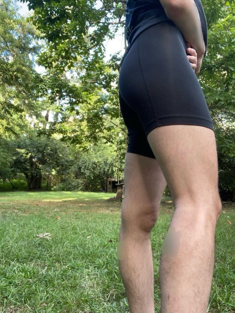 Cedar stands in a clearing in the woods, the photo captures their butt, thighs, and calves with their hands crossed over their abdomen. They wear a black shirt and tight, black lycra shorts. They’re very toned & have soft, blonde body hair. Their tattoos are blurred for privacy.