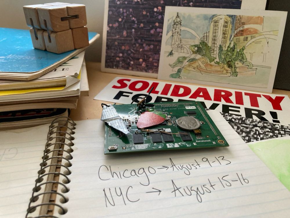 A desktop with an open spiral-bound notebook on it that reads Chicago August 9 through 13, NYC August 15 through 16. A watercolor postcard of Logan Circle, a sticker that says SOLIDARITY FOREVER, a chipboard sculpture in progress, a stack of sheet music notebooks, and a wooden fidget cube are also present 