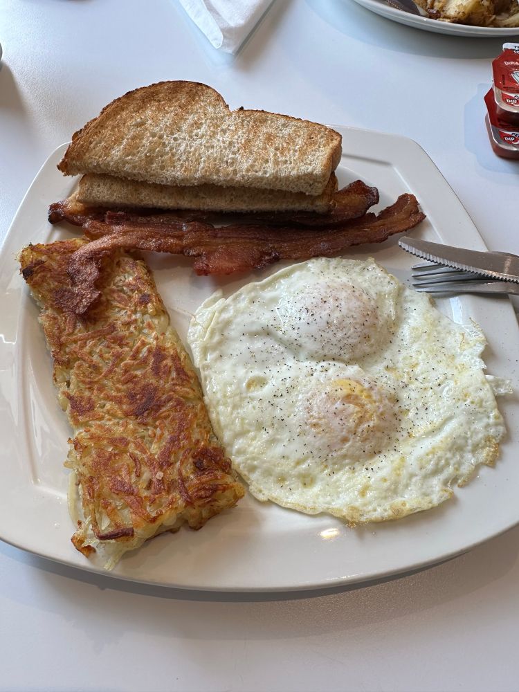 A white plate on a white tablecloth featuring bacon, wheat toast, 2 over medium eggs, and hashbrowns 