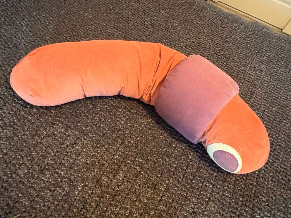 A body pillow with a pink corduroy cover that has a plush purple corduroy collar and large round eyes on one end, to look like a cartoon worm.