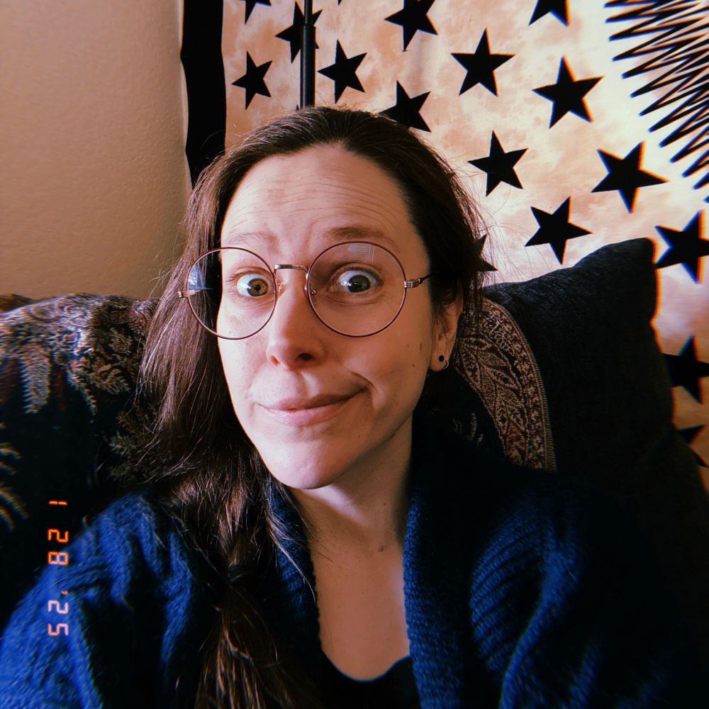 A white woman with long brown hair sitting in a cushioned chair making an unserious face. The metal frames of the glasses being worn are perfectly round and cartoonishly big.
