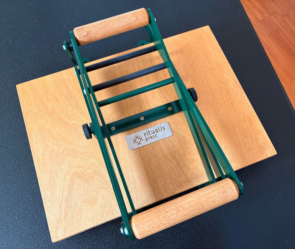 top down view of a closed tabletop wooden printing press with two green arms ending in round wooden handles. A metal plate reads “ritualis press” with a glyph-like logo.