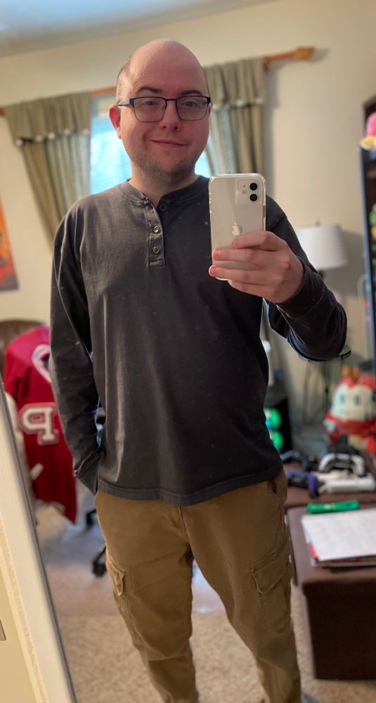 A gay geeky guy in glasses, navy long sleeve Henley shirt, and brown cargo pants smiling at the camera as he stands in this mirror selfie 