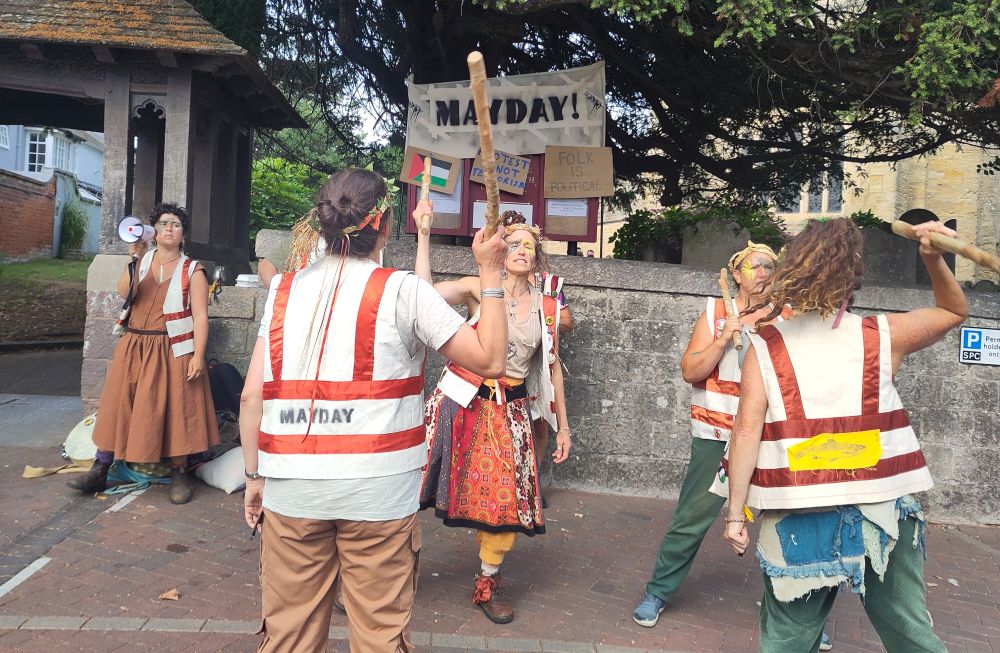 Morris dancers on the street in Sidmouth. One has a print of a salmon pinned to their back. Posters behind them read: Mayday!, Protest is not Terrorism, and Folk is Political.
