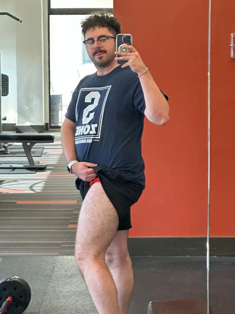 gym selfie of me in a blue shirt and black shorts, but my right short is lifted up to show off my thigh and underwear a bit
