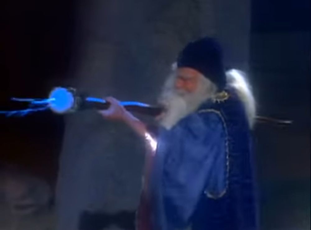 Norman Lloyd as Merlin, in a blue wizard robe and cap and hefting a big wizard staff with a glowing blue end while there is a not real Stonehenge visible behind him. From the Twilight Zone segment “The Last Defender of Camelot.”