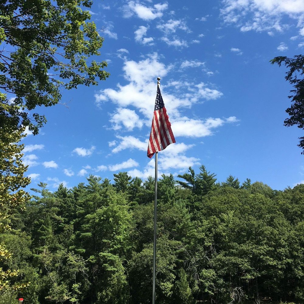 A flagpole with an American flag stands against a blue sky with puffy white clouds and trees in the background.