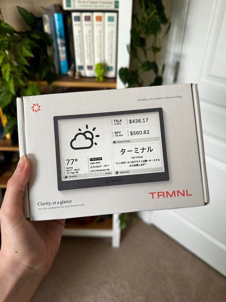 Holding a TRMNL box in front of a bookcase. The TRMNL box shows an e-ink screen with weather and stock ticker widgets.
