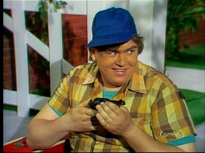 A young John Candy dressed as the Beaver from Leave it to Beaver holding a pistol.