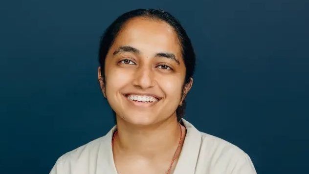 Picture of a brown-skinned person starting to smile at the camera, their black and brown hair is tied on the back. They are wearing a cream shirt, and are standing against a blue background.