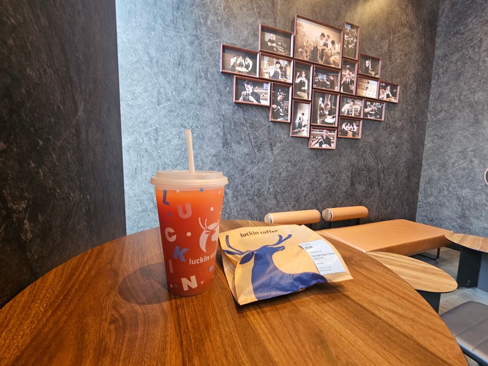 Orange drink in a cup that says Luckin Coffee next to a brown bag with a blue deer like figure on it. Both are on a table. 