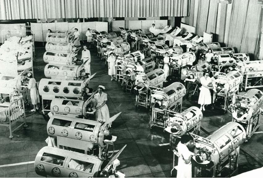Black and white photo of large room filled with iron lungs and patients in them, nurses walking about caring for the patients