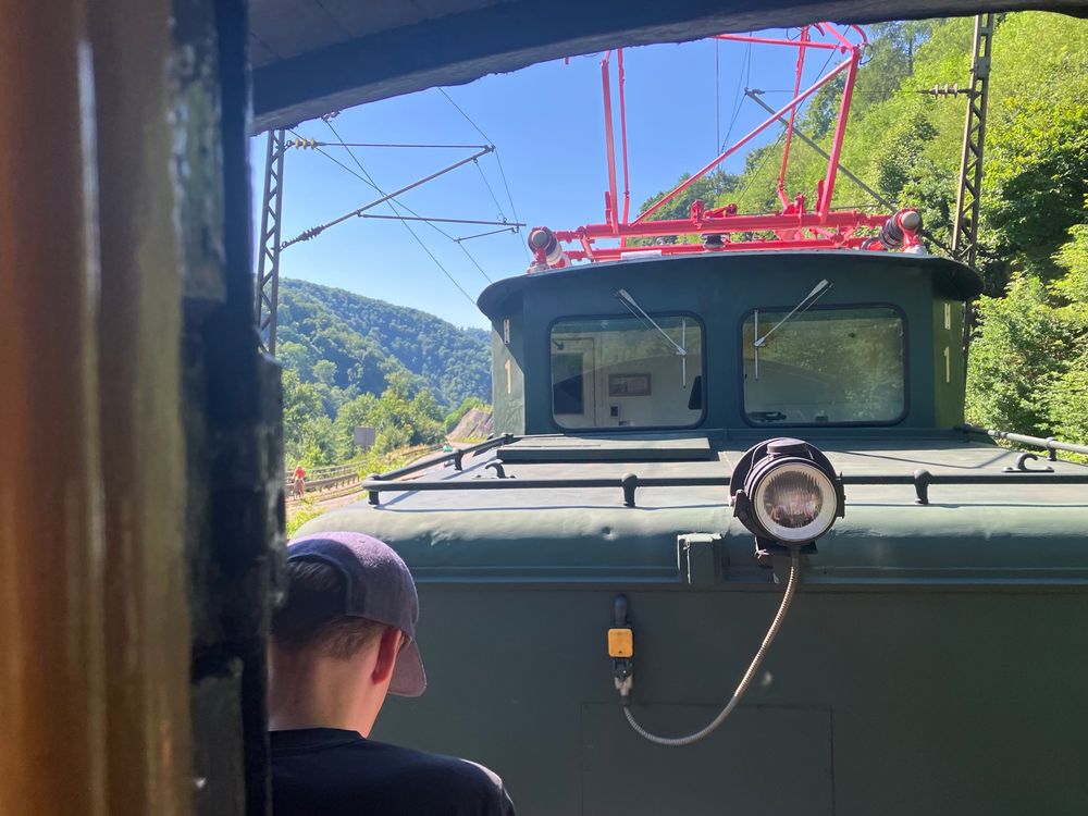 Blick von der offenen Plattform eines Waggons auf eine historische elektrische Lokomotive vom Typ „schwäbisches Krokodil“. Der Zug fährt ins Tal Richtung Geislingen hinab. Links und rechts sind bewaldete Hänge zu erkennen.