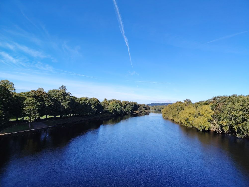 River with clear, almost cloudless blue sky above. The riverbanks are lined with green-leaved trees on both sides (taken late September).