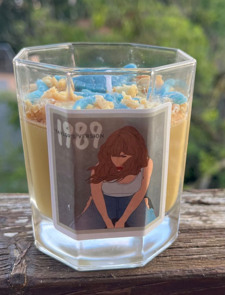 the candle from the side 
the wax is light brown, on top are some wax crumbles in light brown and a star, a tortoise and two mermaid fins in blue. on the glass is a sticker with Taylor sitting on the beach with a white shirt and blue pants and 1989 is written on it 