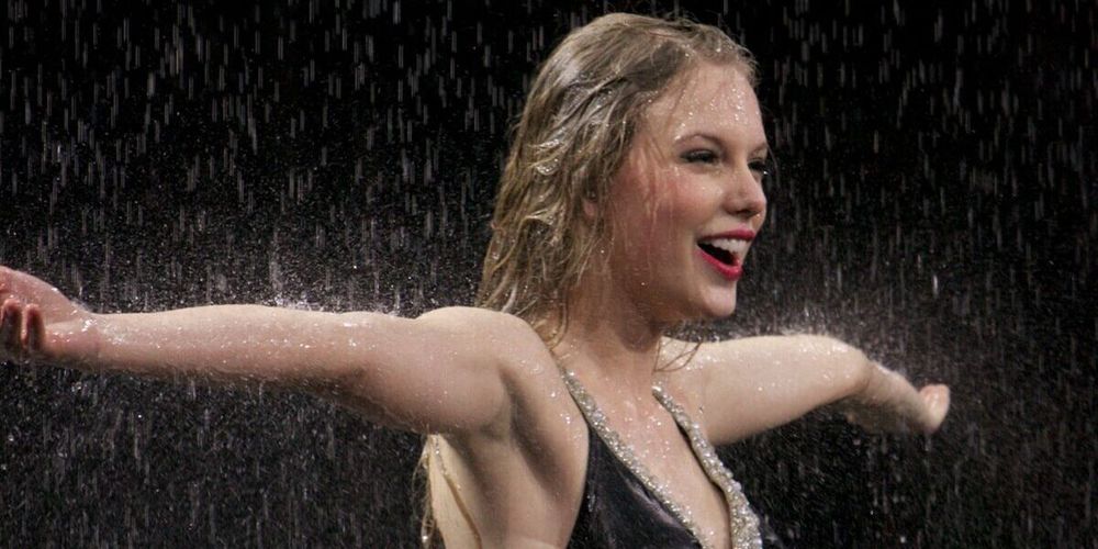 Taylor Swift performing on stage in the rain, smiling with arms outstretched in a wet black dress 