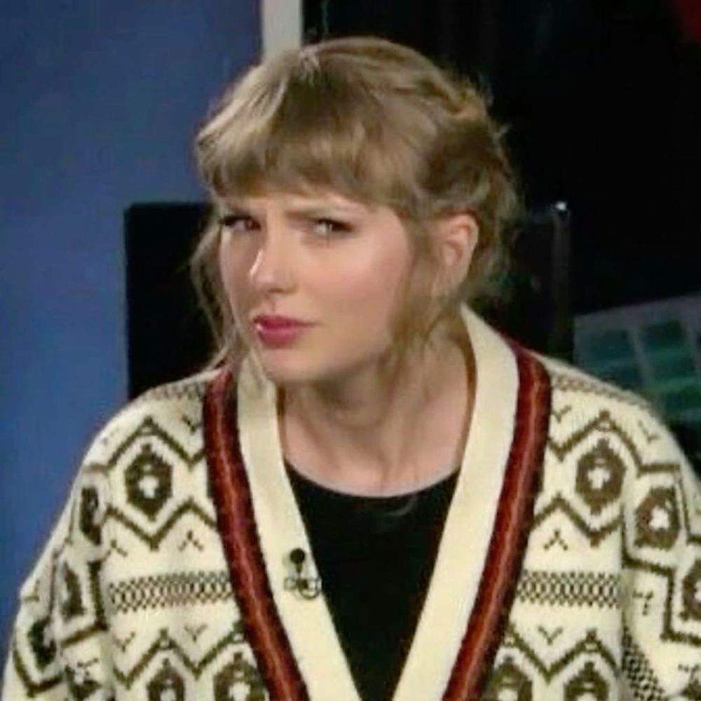 A photo of Taylor Swift, wearing a patterned cream cardigan with red and black accents, looking sideways with a skeptical expression