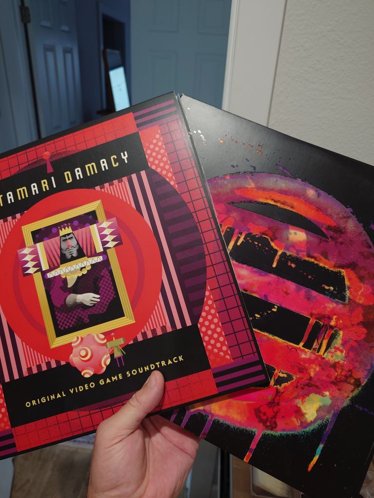 My hand holding two LP albums. On the left is the soundtrack for Katamari Damacy in a bright red with purple and black accents and block letters spelling out the game name in yellow and white text. On the right is the soundtrack for Hotline Miami, on a black background is the game's logo/iconography a circle with three horizontal stripes in a mix of reds and purples in a graffiti style 