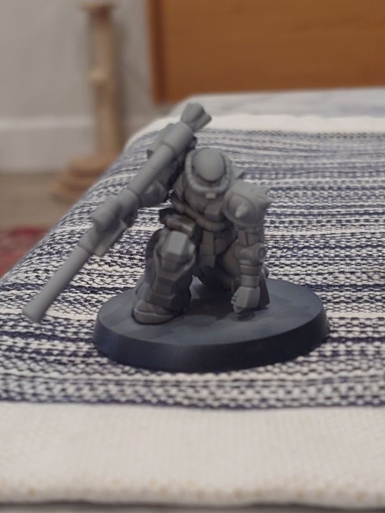 Zaku II mini from the Gundam Assemble line. It is in a crouched pose on one knee with a large bazooka being held. It is covered entirely in grey primer. 