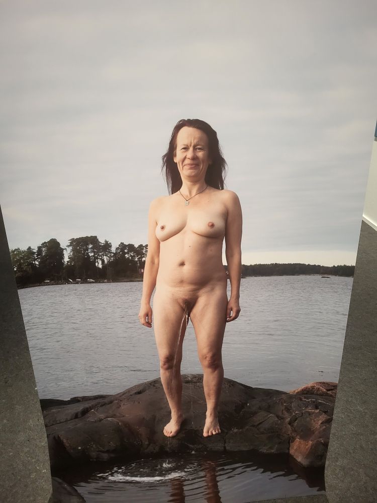 Valokuva alastomasta naisesta, joka seisoo rantakalliolla ja pissaa veteen.