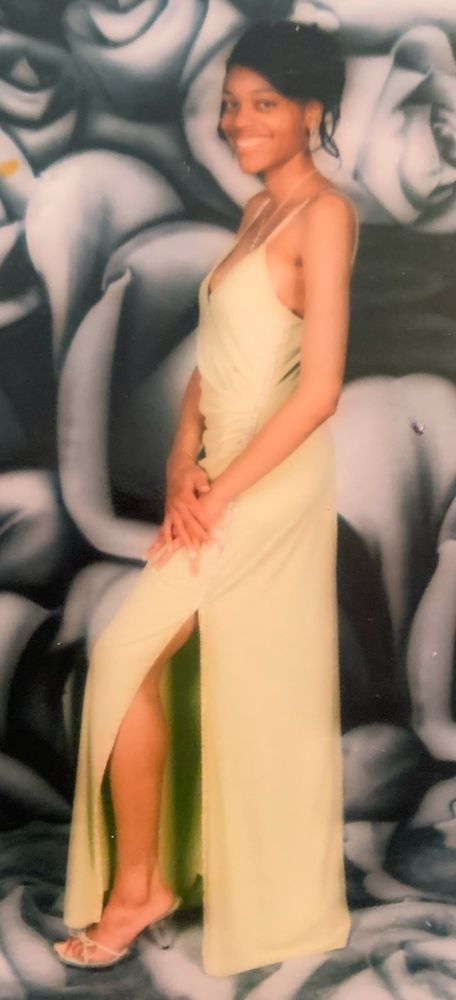 A smiling Black woman in lime green dress with slit to thigh, clear heels, and a gray rose background 