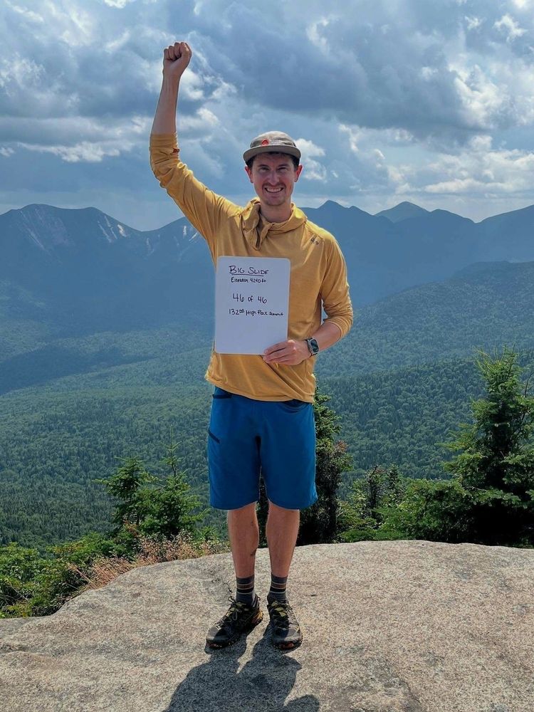 A person stands triumphant on a rock with one arm raised holding a sign in mountainous terrain. The sign reads Big Slide Elevation 4240 ft 46 of 46 13228 High Peak Summit.