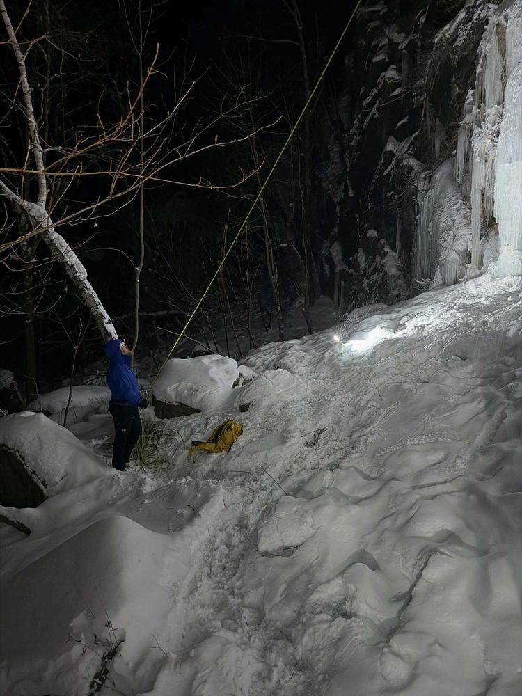 A person in a blue jacket stands holding a rope, surrounded by snow and ice, near a rocky cliff at night. A yellow backpack rests nearby on the snow-covered ground.