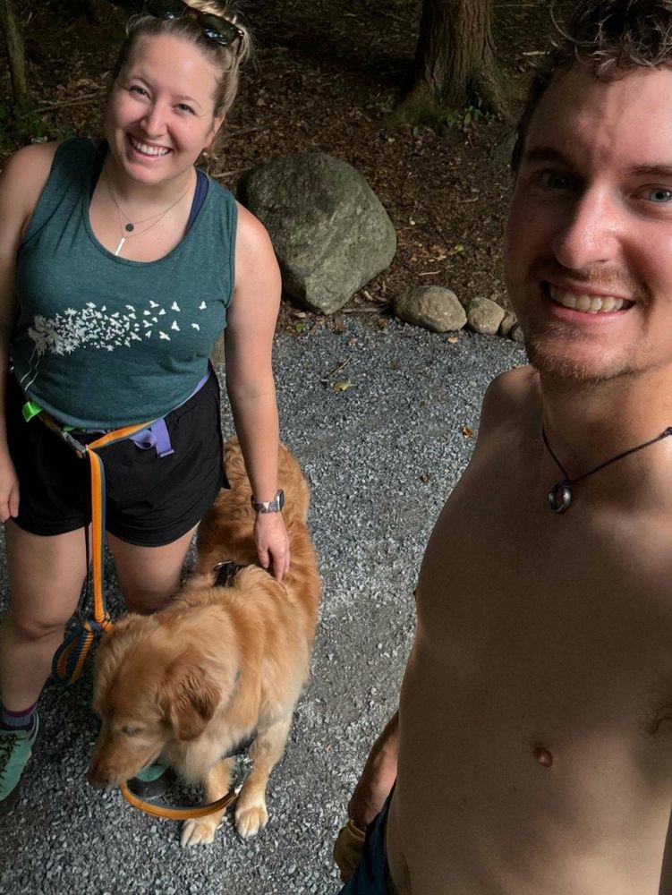 A golden dog stands calmly on a pebble path while two people smile, with trees and rocks in the background.