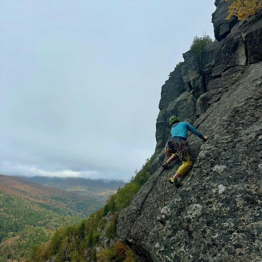 A climber scales a rugged rock face wearing climbing gear and a helmet surrounded by a scenic mountainous vista under a cloudy sky.